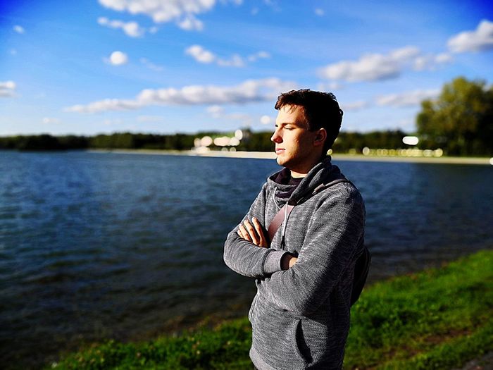 Young man looking away while standing by lake against sky