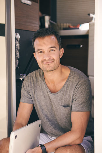 Portrait of mature man holding laptop while sitting on doorway of motor home