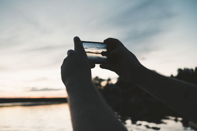 Cropped image of man photographing lake through smart phone during sunset