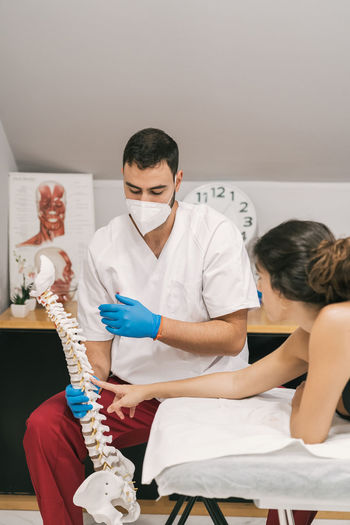 Male physiotherapist explaining spine structure for female patient while using plastic model and sitting in medical room during rehabilitation session
