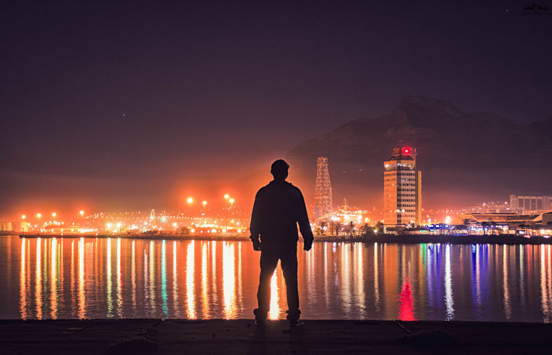 Silhouette man looking at view while standing by sea in illuminated city at night