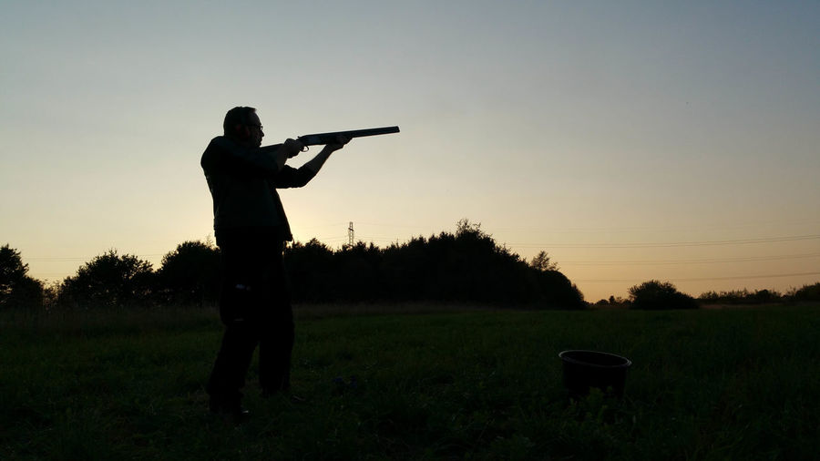 Silhouette man aiming with shotgun on field against clear sky during sunset