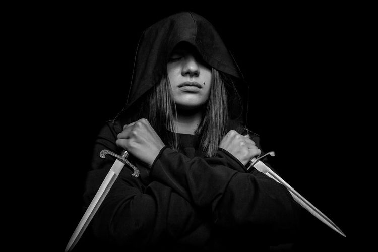 Young woman wearing hood holding swords against black background