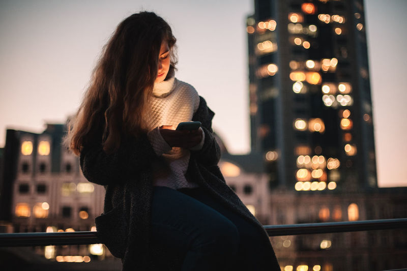 Teenage girl using smart phone sitting on railing in city at night