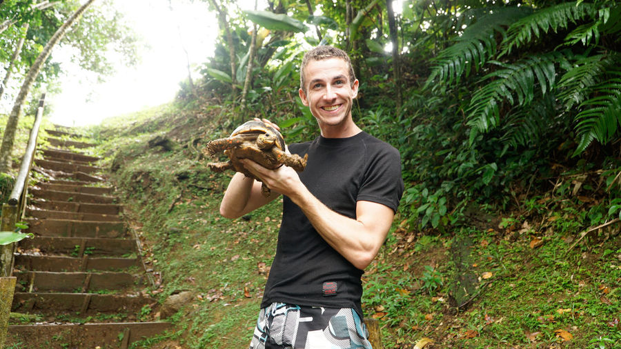 Portrait of smiling young man holding turtle against trees
