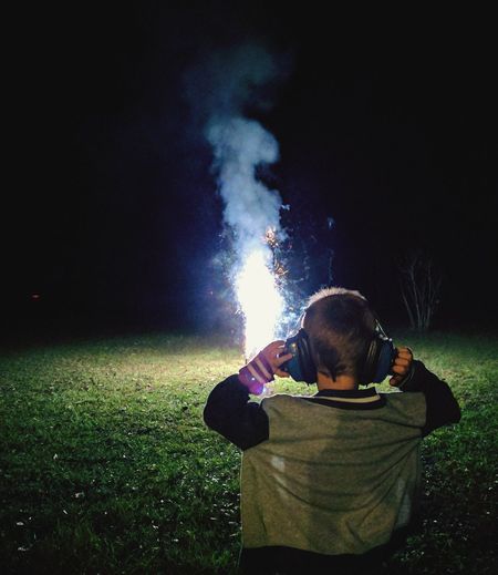 Rear view of boy wearing ear protectors standing on field with firework at night