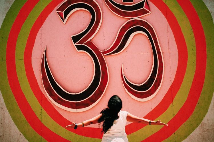 Rear view of woman with arms outstretched standing in front of om symbol on wall