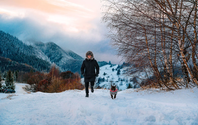 Smiling man and french bulldog dog running in the snow at mountain