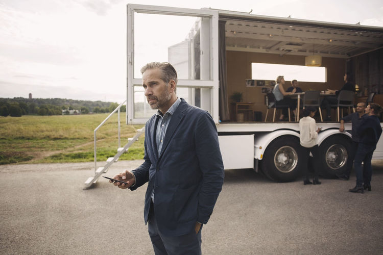 Businessman holding mobile phone with colleagues and portable office truck in background