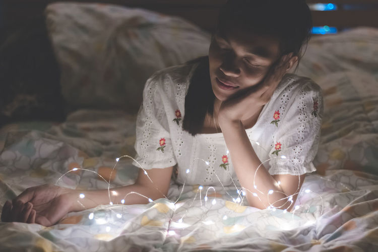 Close-up of young woman lying by illuminated string lights on bed