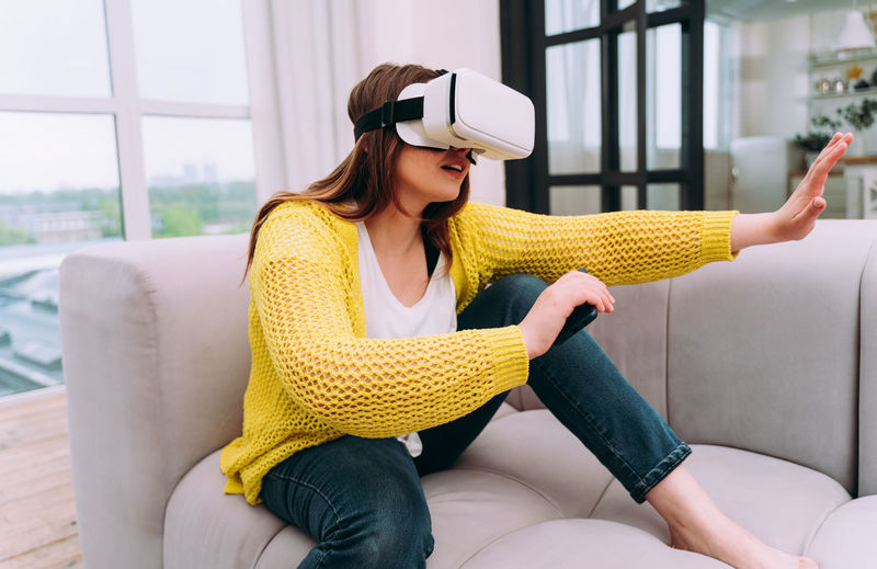 Young woman wearing virtual reality simulator while sitting on sofa at home