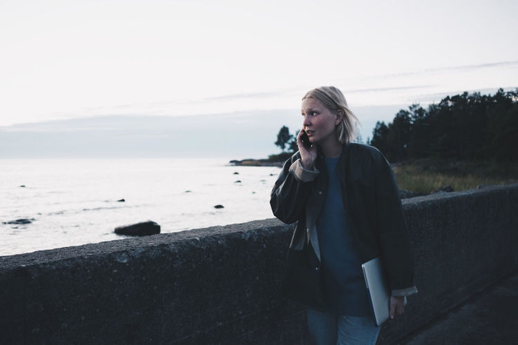 Woman talking on mobile phone and holding laptop while walking by lake against sky