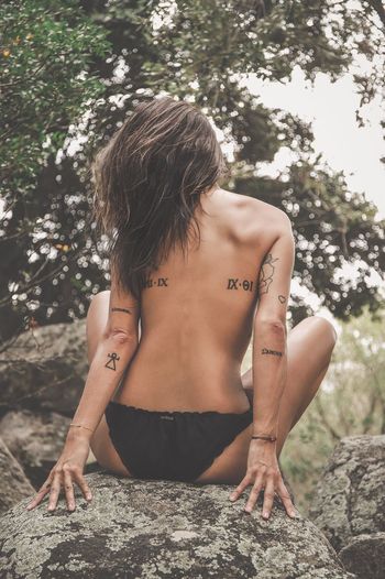 Rear view of topless woman sitting on rock in forest