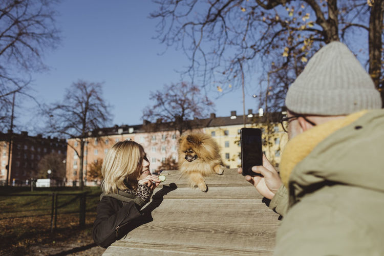 Man photographing woman holding food by pomeranian on obstacle course at park