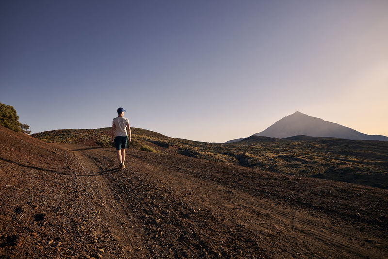 Rear view of man walking on dirt road against beautiful volcanic landscape at sunset. 