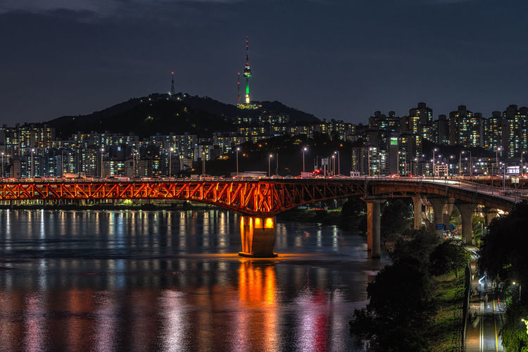 Illuminated bridge over river against buildings at night