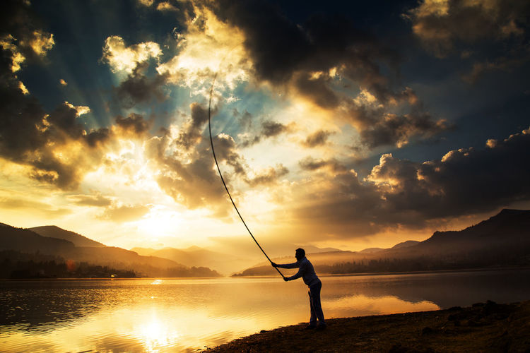 Silhouette man fishing in lake against sky during sunrise