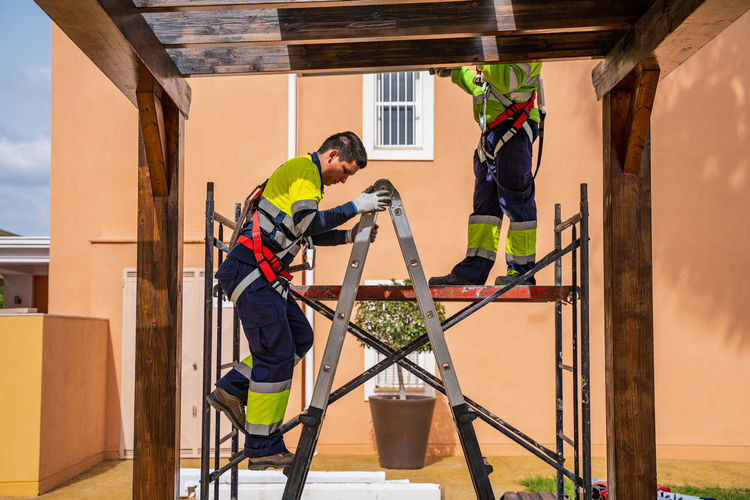 Group of workers in uniform and helmets installing photovoltaic panels on roof of wooden construction near house