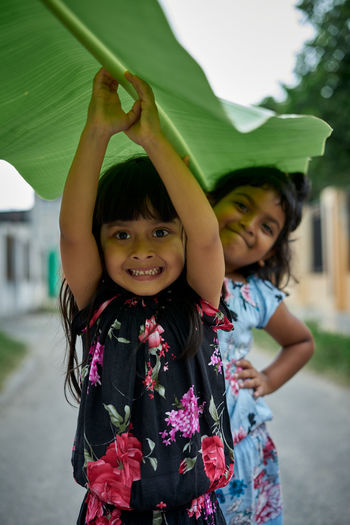 Portrait of smiling girl with arms raised holding banana leaf by sister