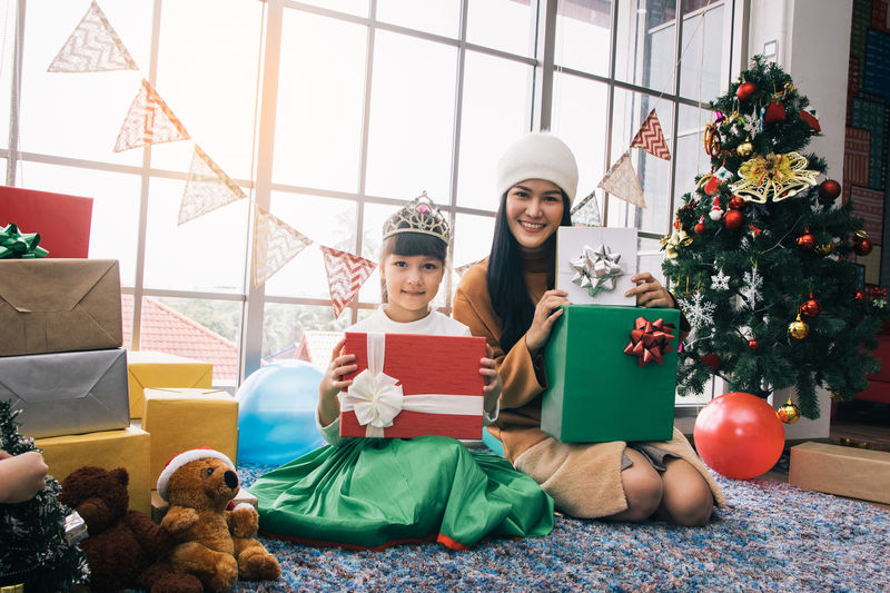Portrait of mother and daughter with gift boxes sitting at home during christmas tree