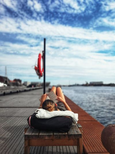 Woman reading book while lying at harbor against sky