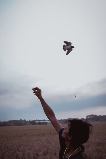 Man with arms raised while bird flying on field against sky