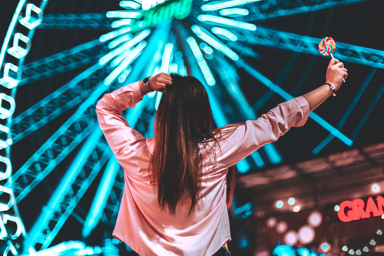 Rear view of woman standing at illuminated amusement park during night