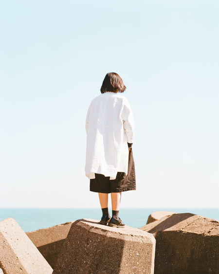Rear view of woman standing on rocks by sea against clear sky