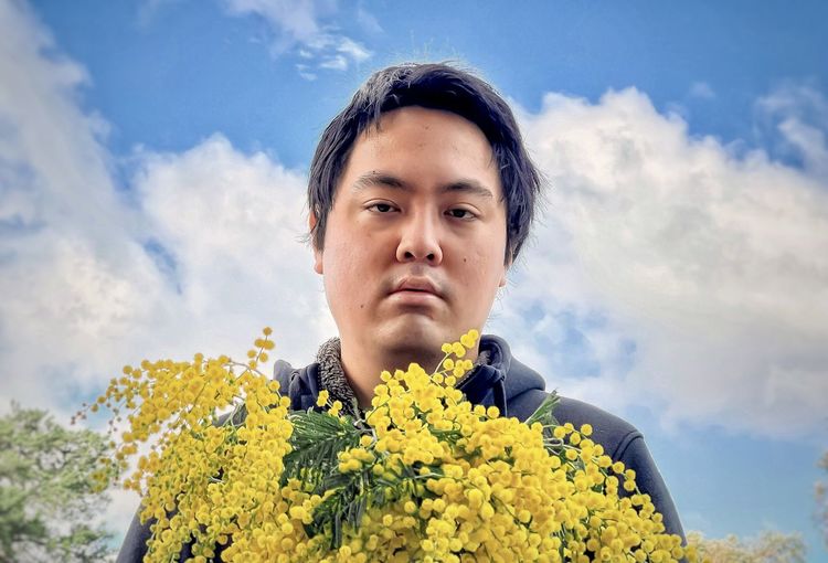 Portrait of young man holding golden wattle flowers against cloudy blue sky.