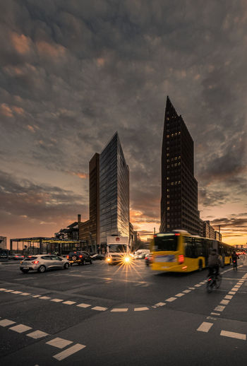 Traffic on road by buildings against sky during sunset