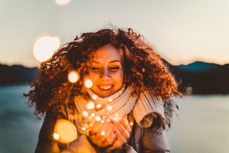 Close-up of smiling woman holding illuminated lights against sky during sunset