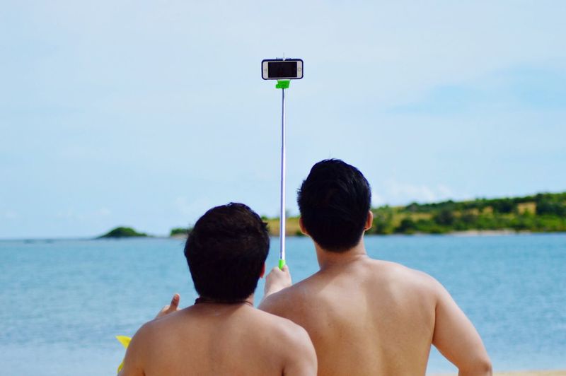 Rear view of shirtless friends taking selfie at beach against sky