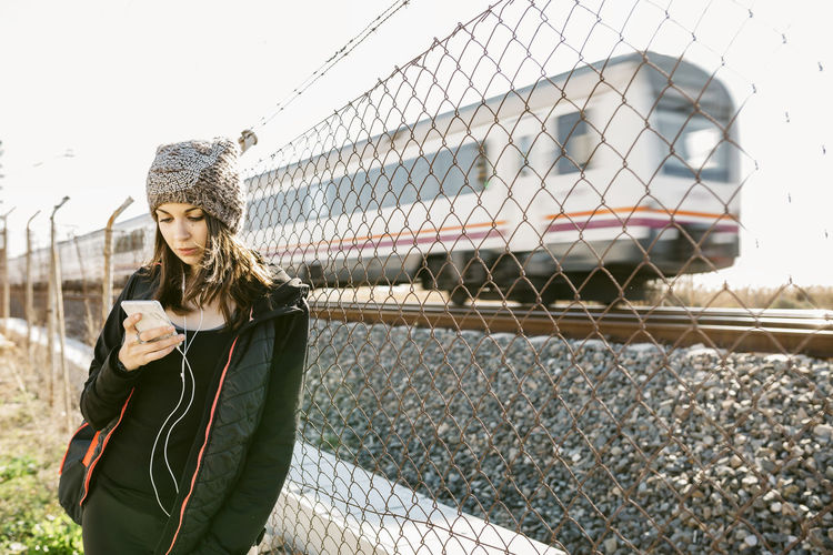 Woman using phone while standing by chainlink fence against train