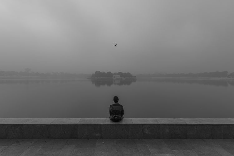 Rear view of person sitting on retaining wall by kankaria lake during foggy weather