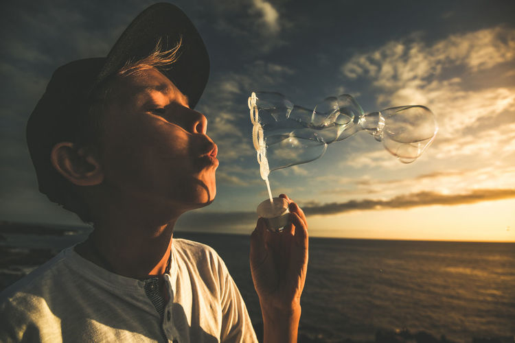 Close-up of cute boy blowing bubbles against sky during sunset