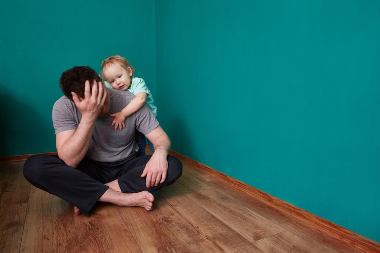 The child feels sorry for the upset father sitting on the floor in a gloomy green room.