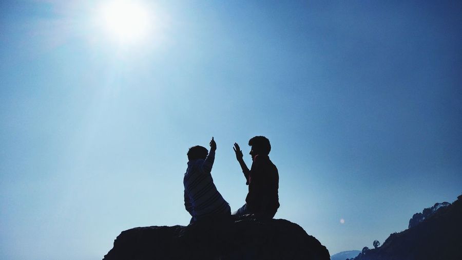 Men doing high-five while sitting on rock against clear sky during sunny day