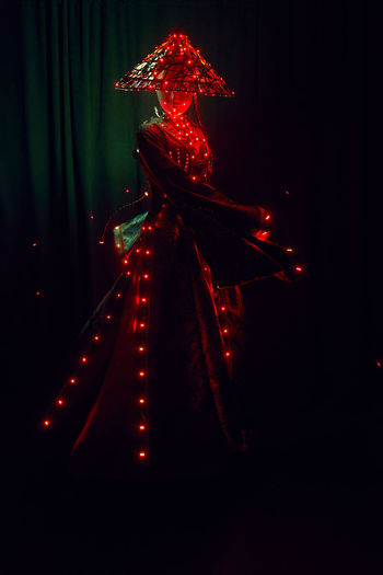 Unrecognizable female with a mask in creative traditional outfit and vietnamese headwear with red illumination standing in dark studio on black background during performance