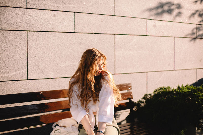 Portrait of young woman sitting on bench by wall in city in summer