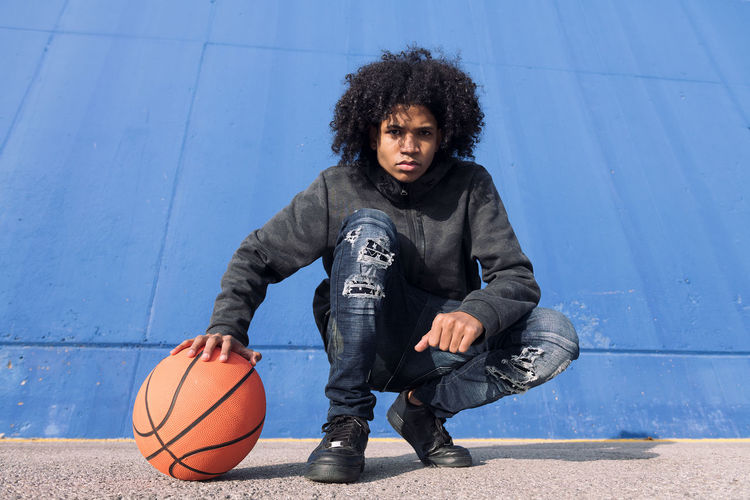 Full body of serious african american teen male in informal outfit sitting with basketball ball near blue wall and looking at camera