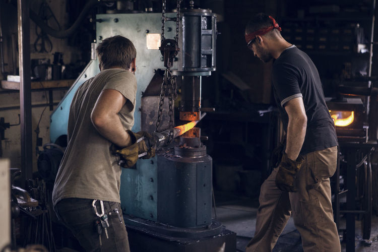 Blacksmith standing by coworker forging rod in machinery at workshop
