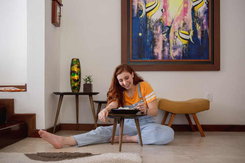 Full body barefoot young woman smiling and eating sushi with chopsticks while sitting on floor in stylish room at home