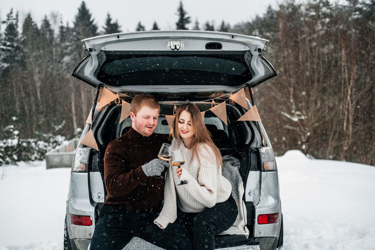 Couple holding wineglass while sitting by car trunk during winter