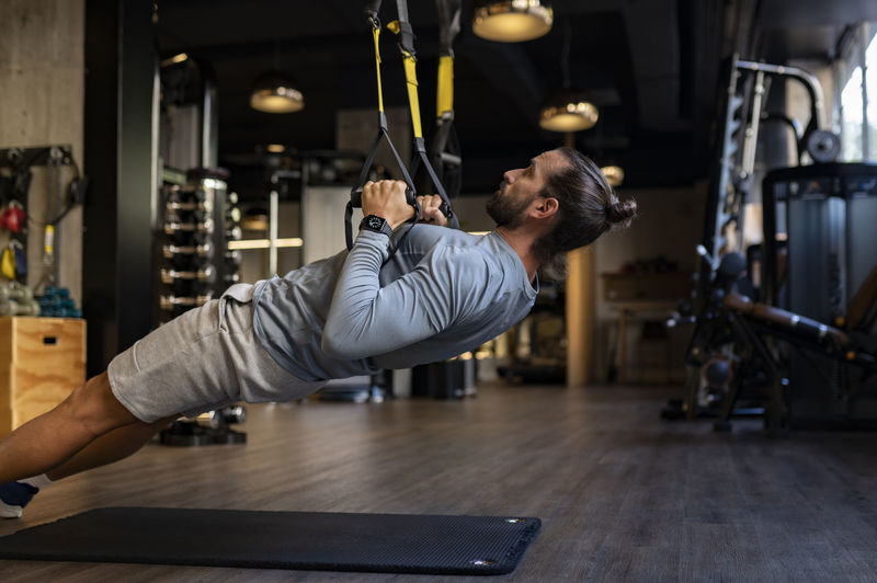 Side view of concentrated muscular male athlete practicing suspension training with trx ropes in modern gym