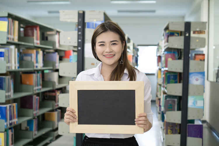Portrait of smiling young woman holding writing slate in library