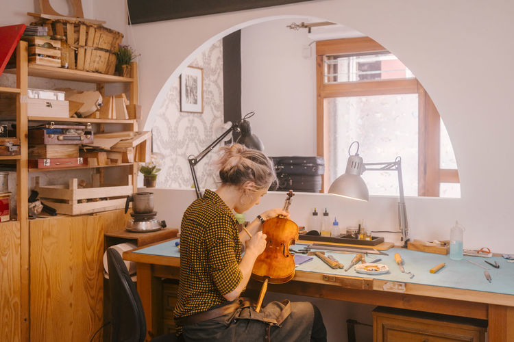 Instrument maker with paintbrush polishing violin at desk in workshop