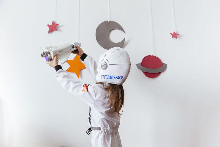 Girl wearing space suit playing with rocket against wall at home