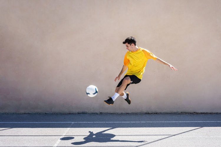 Young male player kicking football while practicing soccer against building