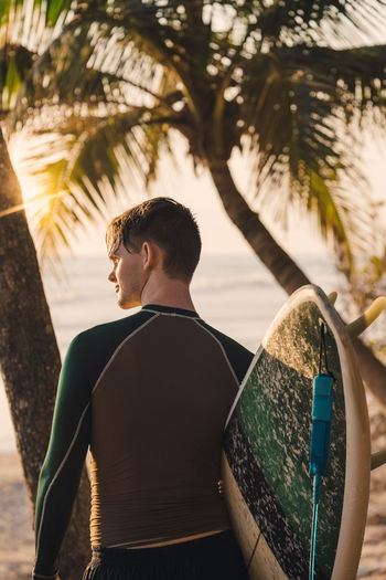 Rear view of young male surfer with surfboard at beach during sunset