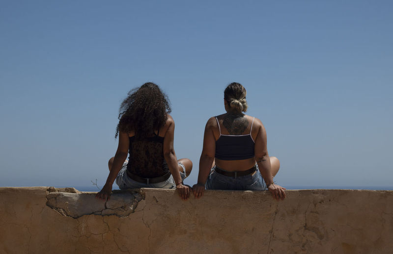 Rear view of two women sitting on wall beside the sea against clear blue sky